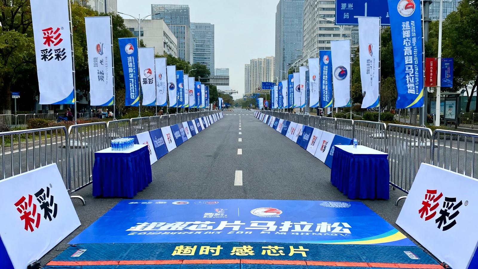 一场城市马拉松赛事的起终点区域，彩神提供的铁马围栏、计时芯片感应垫、补给站桌台和品牌道旗整齐布置完毕